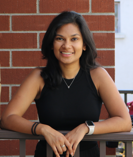 A picture of Anisha smiling in a black top, in front of a brick wall. She has shoulder length black hair and a necklace.